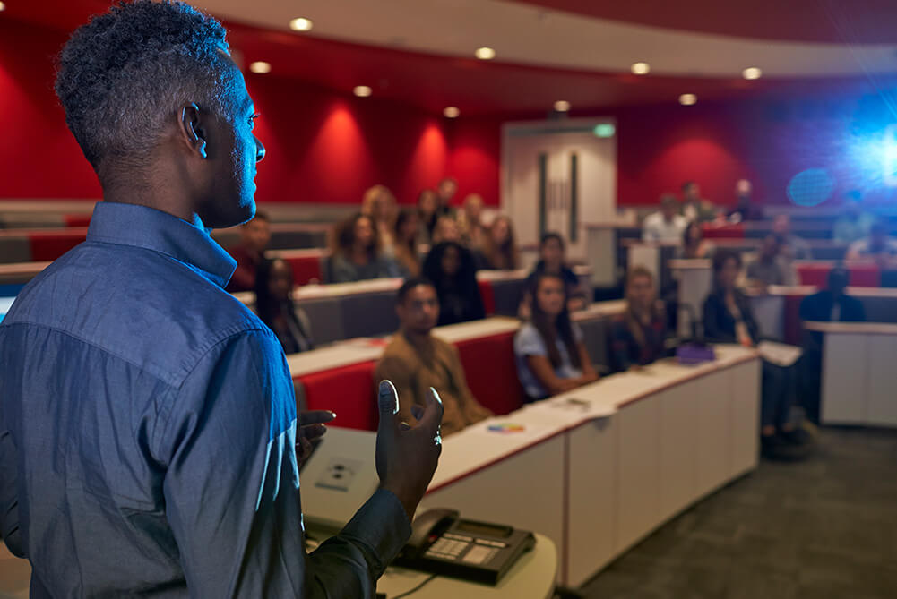 A black man giving a public speech to a class of students sitting in a conference hall with red walls