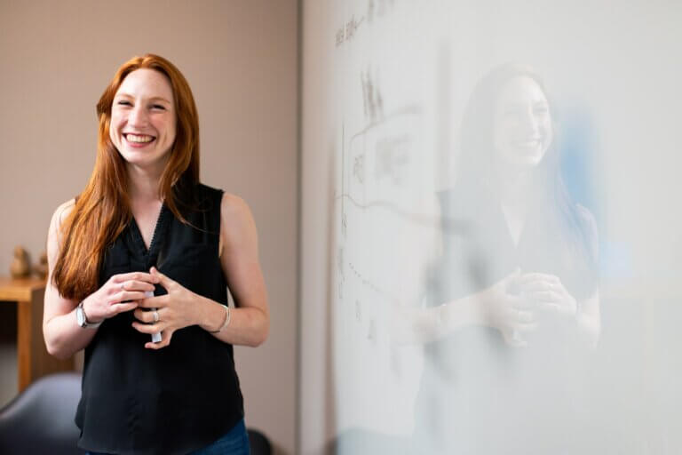 Smiling girl with red hair, dressed in a black blouse, standing beside a whiteboard.