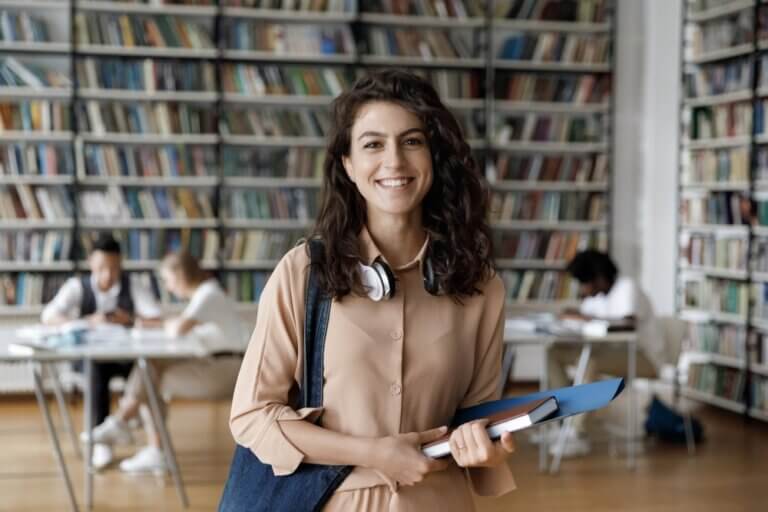 smiling woman studying to be a doctor of philosophy, holding books and standing inside a library